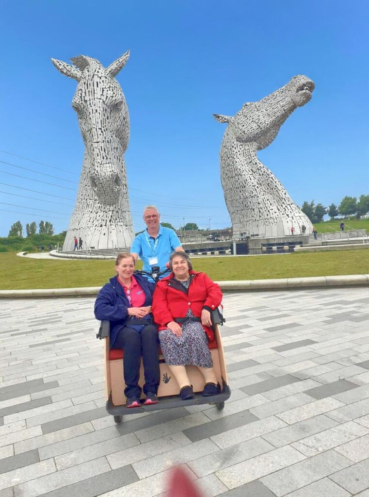 A carer from Creative Homecare poses with a customer in front of the Kelpies sculptures in Falkirk on a sunny day, with clear blue skies and green grass in the background.